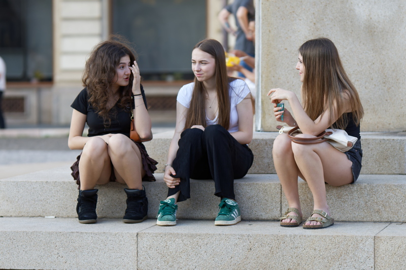 Three young women sitting on concrete stairs outdoors, dressed in casual summer outfits and being engaged in a friendly conversation